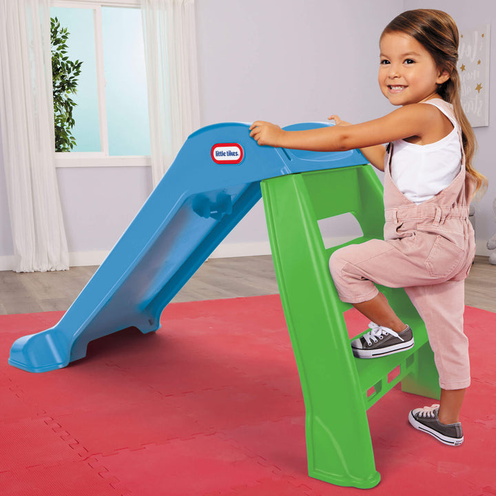 Child standing on a green and blue slide with a Little Tikes logo indoors.