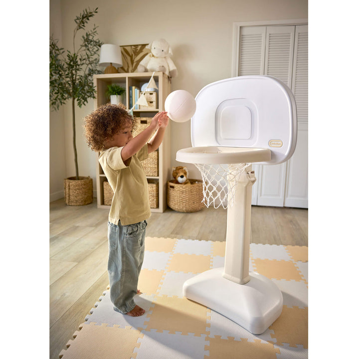 Child with brown curly hair, tan shirt and blue jeans tossing a ball into the hoop. 