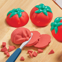 Red tomato-shaped play sand with green leaves on a wooden surface, accompanied by a blue tool.