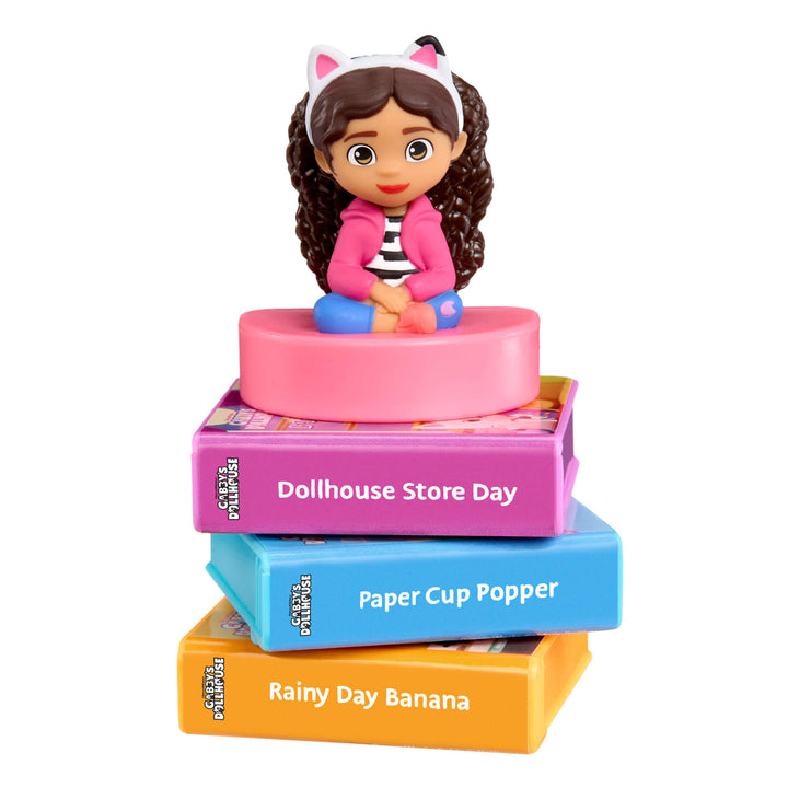 Toy figure of a girl with a pink dress on top of books labeled 'Dollhouse Store Day', 'Paper Cup Popper', and 'Rainy Day Banana' on a white background.