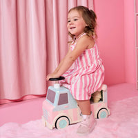 A young girl with blonde hair in a pink and white striped dress sits on a pastel pink and light blue toy truck in a pink room with a pink curtain backdrop.