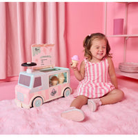 A young child wearing a pink and white striped dress sits on a fluffy pink rug next to a toy ice cream truck, holding a toy ice cream cone in a pink room.
