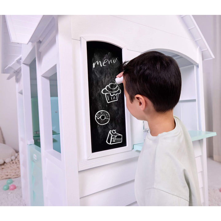 Child writing on a chalk board attached to the outside of a white playhouse.