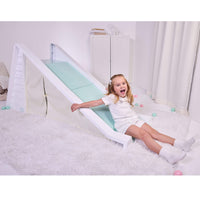 Girl with long blond hair dresses in white sitting at the bottom of a white and seafoam green slide with a play tent area underneath. Slide is on a white shag rug