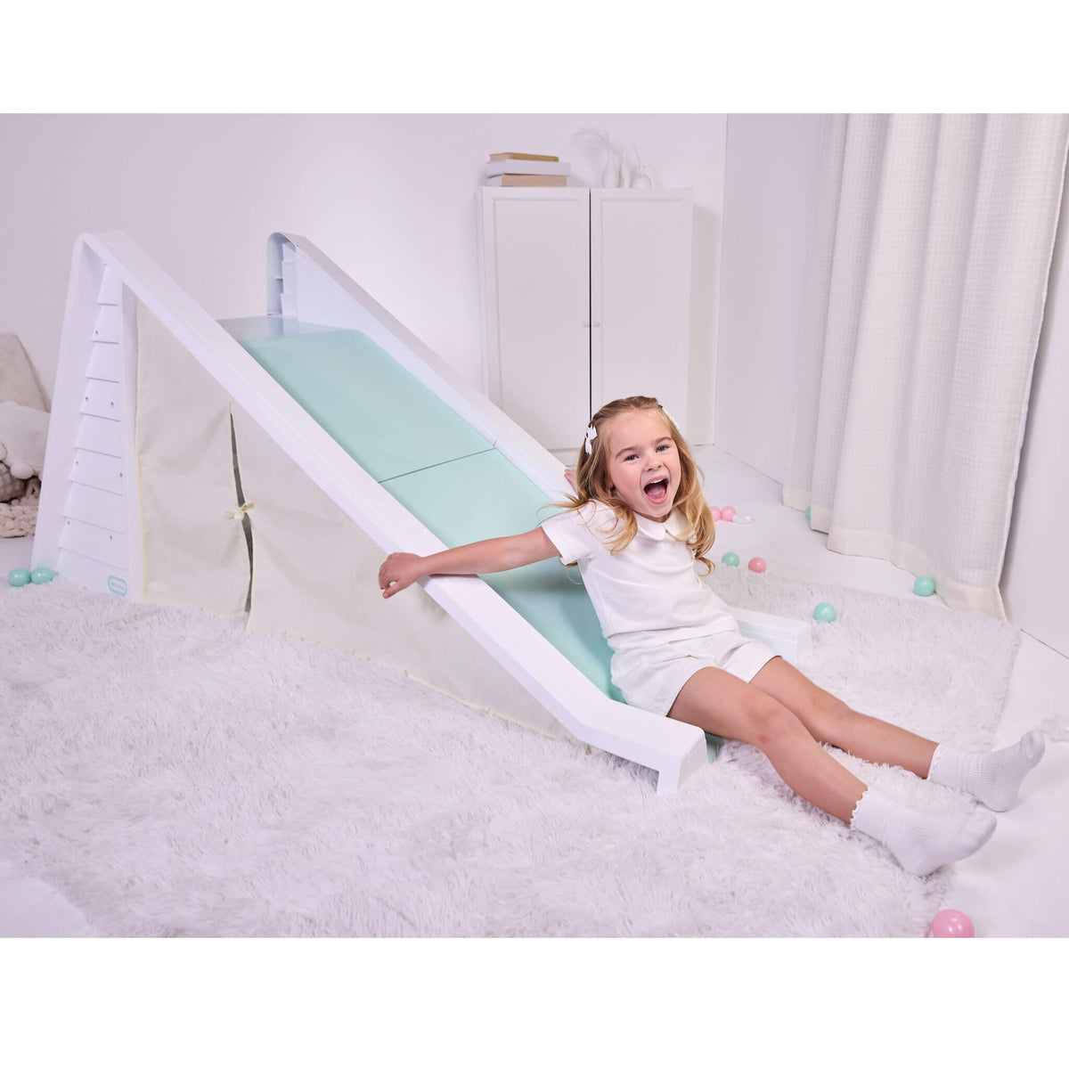Girl with long blond hair dresses in white sitting at the bottom of a white and seafoam green slide with a play tent area underneath. Slide is on a white shag rug