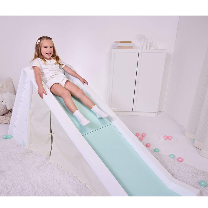 Girl with long blond hair dresses in white sitting at the top of a white and seafoam green slide in a play room setting.