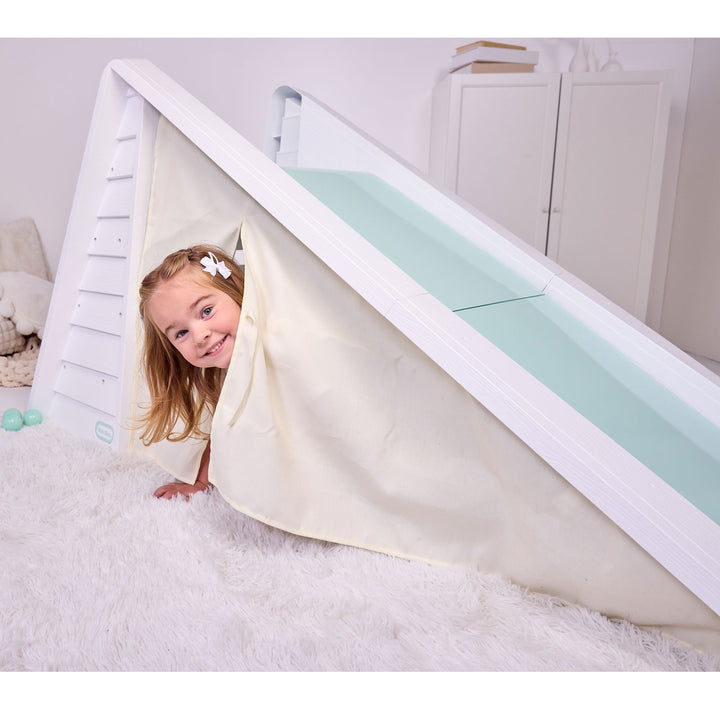 A young child with a white flower hair clip peeks out from inside a 'play tent' under a slide on a white shag rug.