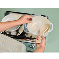 A child's hands holding a translucent plastic saucepan lid and a cream-colored spoon over a toy stovetop, with other white cookware visible in the background.