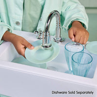 A child in a mint green shirt washes light blue dishes in a white sink with a chrome faucet. Two clear blue glasses and a matching plate are visible in the sink, with the text 'Dishware Sold Separately' at the bottom right.
