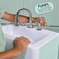 A child's hands and arms are shown interacting with a chrome faucet and handles on a white sink. The background is a mint green paneled wall, and a light blue speech bubble graphic in the top right reads 'PUMPS WATER'.