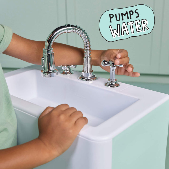 A child's hands and arms are shown interacting with a chrome faucet and handles on a white sink. The background is a mint green paneled wall, and a light blue speech bubble graphic in the top right reads 'PUMPS WATER'.