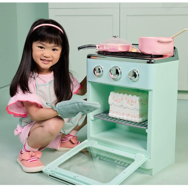 A young girl with long black hair and a pink headband crouches beside a mint green toy stove oven. The toy stove has silver dials, pink pots, and a white-and-pink cake inside, with the oven door open.