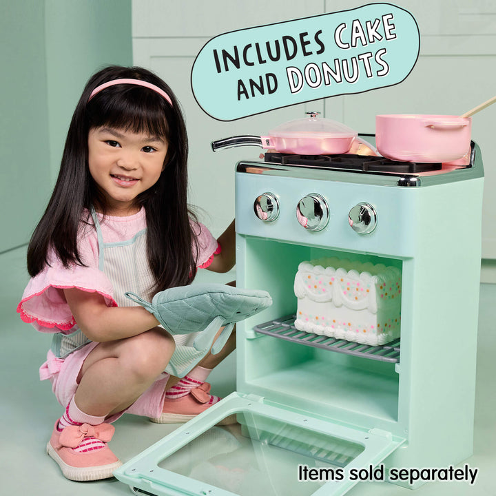 A young girl in a pink outfit kneels beside a mint green toy play stove, holding a gray oven mitt. The toy stove has silver knobs, pink pots, and a white cake on the shelf, with text overlays reading 'INCLUDES CAKE AND DONUTS' and 'Items sold separately'.