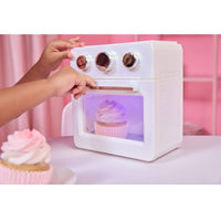 A white toy oven with a transparent door showing a pink cupcake inside, with two hands turning dials on top. A play cupcake sits in the foreground on a pink table against a pink background.