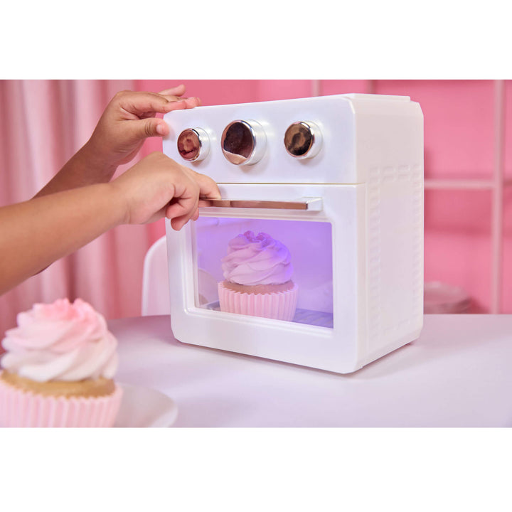 A white toy oven with a transparent door showing a pink cupcake inside, with two hands turning dials on top. A play cupcake sits in the foreground on a pink table against a pink background.