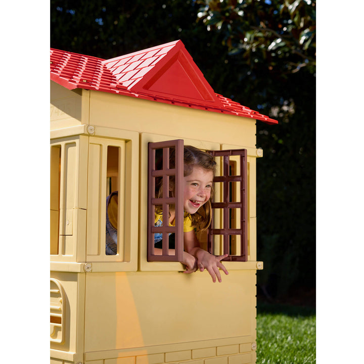 A child smiling and leaning out of an open window of a playhouse with a red roof, set outdoors on grass with trees in the background.