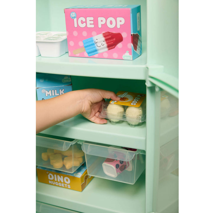 A hand placing 'eggs' into a mint green toy refrigerator. The organizer contains small toy food items, with other toy refrigerator contents visible on the shelves. 