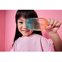 A young girl with dark hair wearing a pink top holds a translucent, ice pop over one eye against a solid pink background.