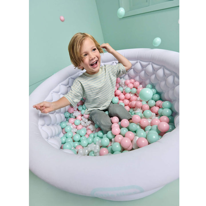 A smiling young child sits inside a large inflatable ball pit filled with pastel pink and mint green plastic balls in a mint green room.