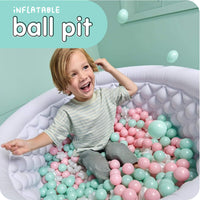 A smiling young child sits inside a white inflatable ball pit filled with pastel pink and mint green plastic balls in a mint green room. A teal graphic overlay in the top left reads 'INFLATABLE ball pit'.