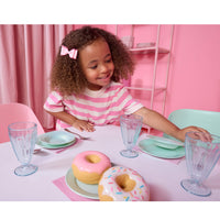 A young girl with curly hair at a pastel seafoam green table setting, smiling near two toy donuts. The scene includes mint green plates, clear glass cups, pink napkins, and a pink chair in the background.