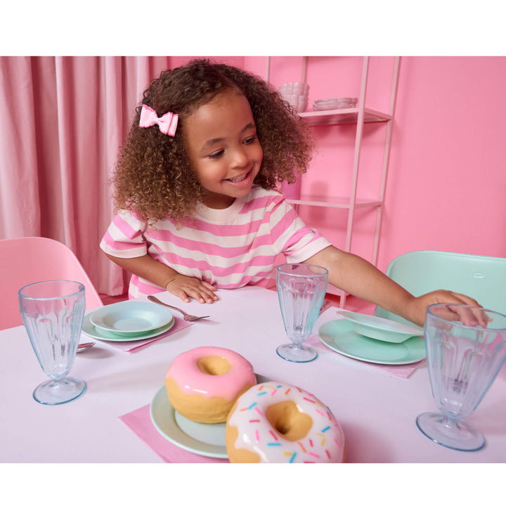 A young girl with curly hair at a pastel seafoam green table setting, smiling near two toy donuts. The scene includes mint green plates, clear glass cups, pink napkins, and a pink chair in the background.