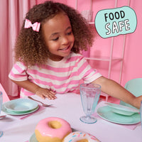 A young girl with curly hair wearing a pink and white striped shirt sits at a table with mint green dishes and donuts in a pink room. A light blue badge in the top right reads 'FOOD SAFE'.