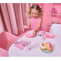A young girl with blonde pigtails at a pink table playing with a pink toy dinner set, holding a pink plate and smiling. The set includes pink plates, glasses, napkins, and toy donuts on a light blue plate.
