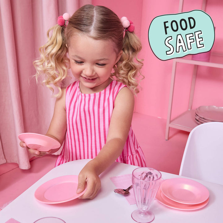 A young girl with blonde pigtails wearing a pink and white striped dress sits at a table playing with pink toy plates and a glass, in a pink room. A light blue speech bubble in the top right says 'FOOD SAFE'.