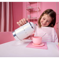 A young girl smiling while pouring water from a white and chrome kettle  into a pink teacup and saucer on a table, set against a pink room background.