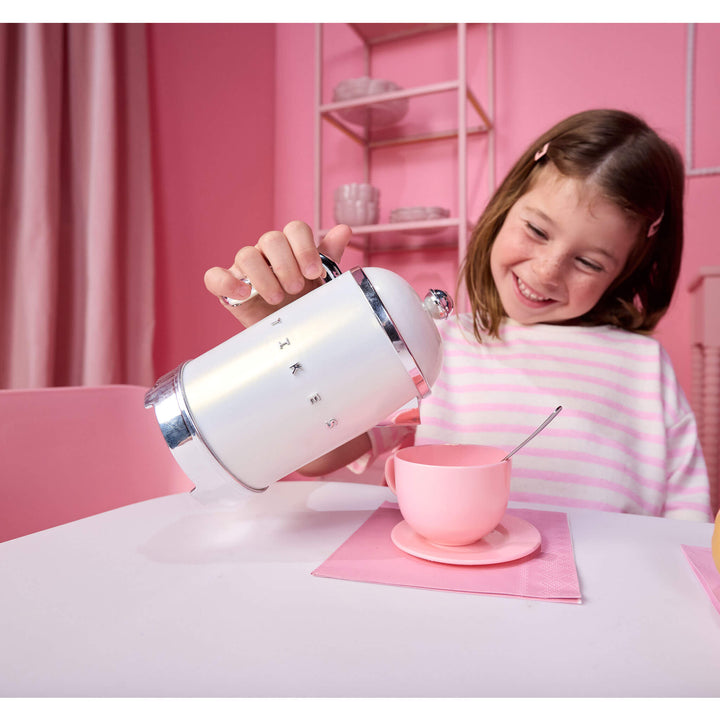 A young girl smiling while pouring water from a white and chrome kettle  into a pink teacup and saucer on a table, set against a pink room background.