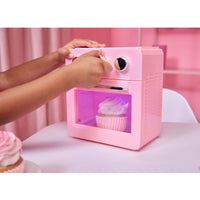A child's hands turn a knob on a small pink toy oven with a cupcake inside, set on a table against a pink background.