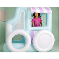 A child stands inside a large inflatable play structure shaped like a light blue and white truck in with pink-and-white striped awning details, photographed in a pastel-colored room.