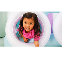 A young girl with long brown hair smiles while crawling inside a white inflatable tunnel with a pink padded interior, set against a light blue and purple play area.