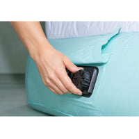 A close-up of a person's hand pressing a black valve or connector onto a light aqua inflatable sleeping pad or bag.