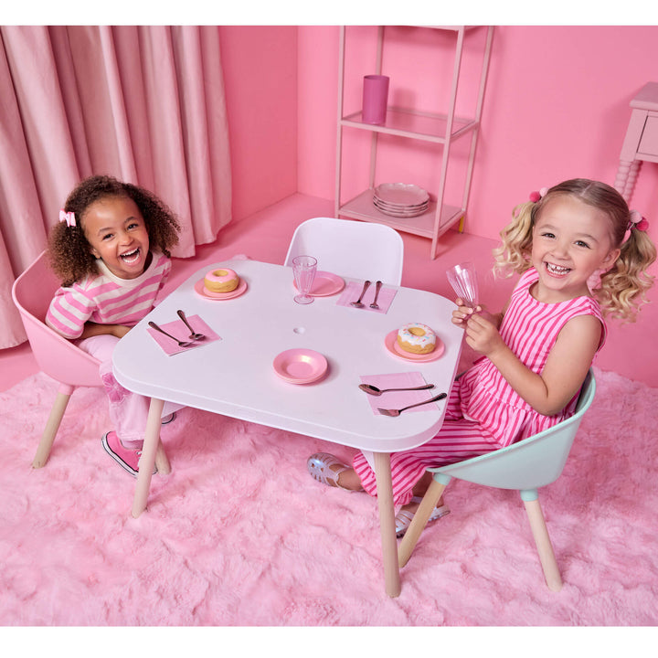 Two young girls sitting at a white toy table with a pink faux fur rug in a pink room. The girl on the left is wearing a pink and white striped shirt and sitting in a pink chair, while the girl on the right is wearing a pink and white striped dress and sitting in a mint green chair.