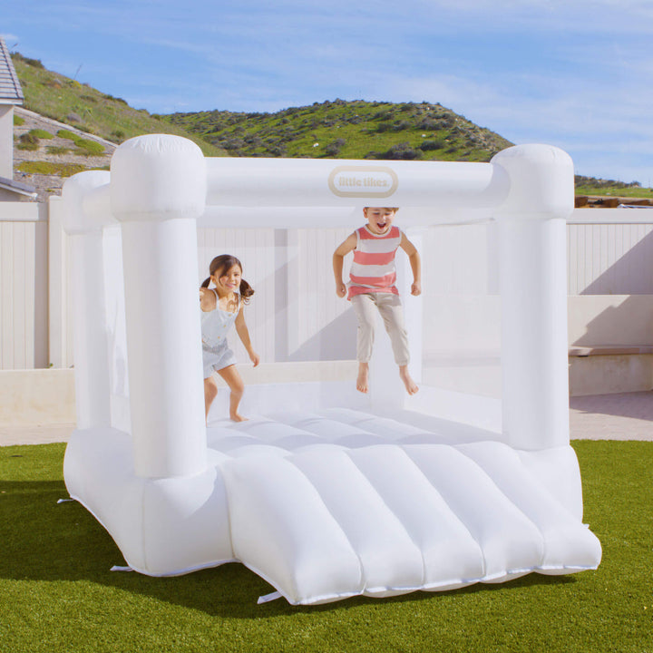 A children's inflatable bounce house set up on a green lawn outdoors, with two kids jumping inside. The product is white and a small visible Little Tikes logo on the top frame.