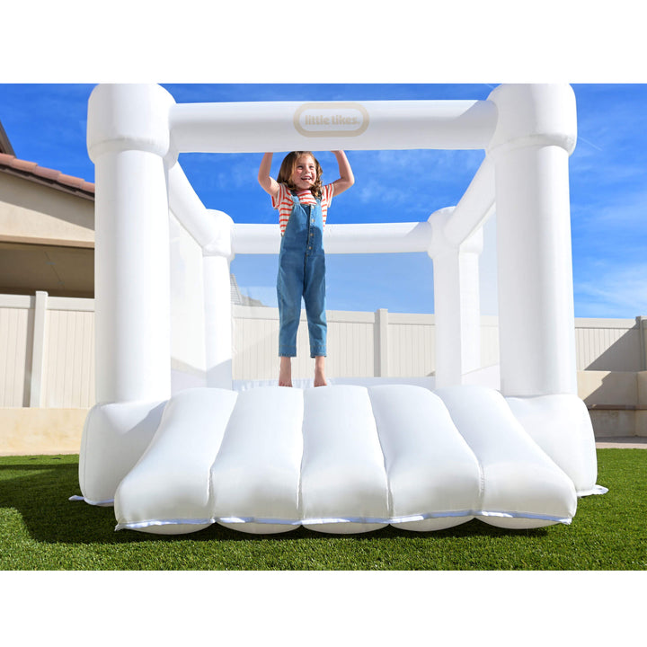 A child stands inside a white inflatable bouncy castle outdoors on grass under a blue sky. The castle has thick padded walls and a slide at the front.