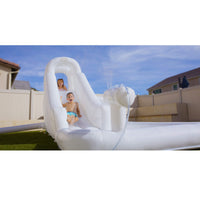 A white inflatable pool slide with water spraying from a hose, with two children sliding down in an outdoor backyard setting under a blue sky.