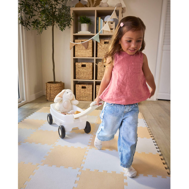 Girl with brown hair, wearing a pink shirt and jeans pulling the wagon inside on a play mat. Fake tree and book shelves in the background.