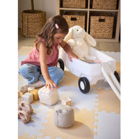 Girl with brown hair, wearing a pink shirt and jeans sitting on the play mat next to the wagon putting toys into it.