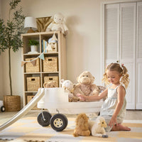 Child using white wagon with rails removed to put stuffed animals in. Indoor setting with beige walls, bookcase and fake tree