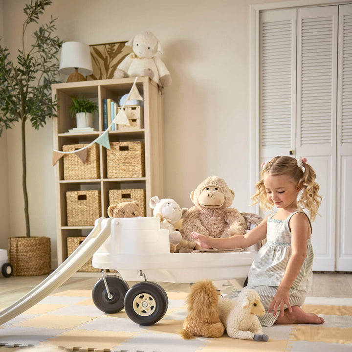 Child using white wagon with rails removed to put stuffed animals in. Indoor setting with beige walls, bookcase and fake tree