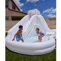 An inflatable outdoor water slide pool with a shallow pool at the base, shown on a lawn with two children playing under a blue sky. 