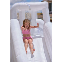 A young girl in a pink swimsuit slides down a white inflatable pool slide outdoors, with water spraying from a small overhead shower feature.