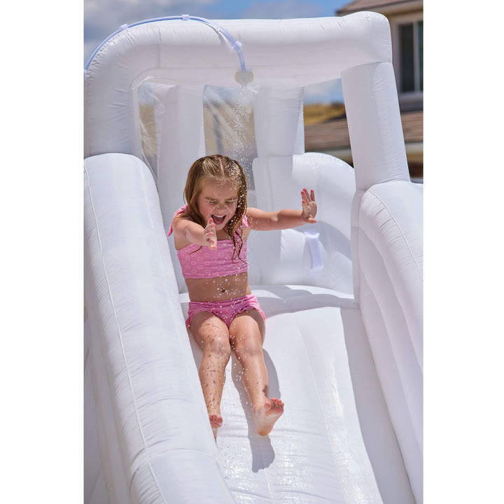 A young girl in a pink swimsuit slides down a white inflatable pool slide outdoors, with water spraying from a small overhead shower feature.