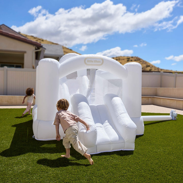 A white Little Tikes inflatable bouncy castle with a slide, set up on green grass in a backyard with two children playing under a blue sky.