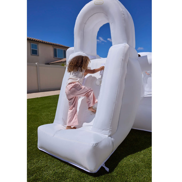 A child in a white shirt and pink pants climbs a white, outdoor inflatable on a green lawn under a blue sky.