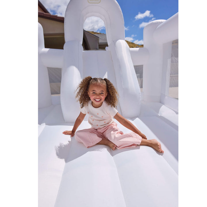 A young girl with curly hair sits smiling on a large white inflatable bouncy castle outdoors under a blue sky.