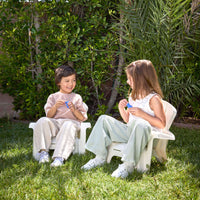 Two kids sitting on the white chairs, on the grass with tree behind them
